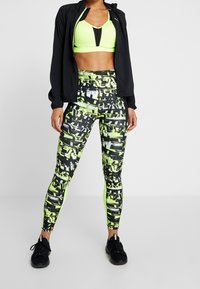 Nainen, jolla on päällään neonkelturneinen urheiluliivi, musta takki, kuvioidut mustat ja neonkelturneiset legginsit sekä mustat urheilukengät, seisoo valkoista taustaa vasten.