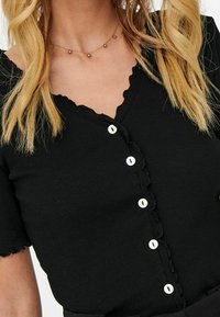 Black ribbed button-up top with short sleeves, scalloped neckline, and white buttons. Features a fitted design and textured fabric.