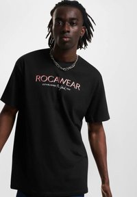 Fekete pamut póló, piros és fehér "ROCAWEAR" logóval, rövid ujjakkal, kerek nyakkal és laza fazonnal. Ezüst lánc nyaklánc látható.