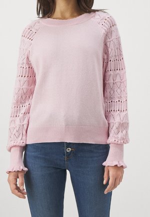 Jasnoróżowy sweter dziergany z wzorzystymi, półprzezroczystymi długimi rękawami i falbanianymi mankietami, noszony z niebieskimi dżinsami.