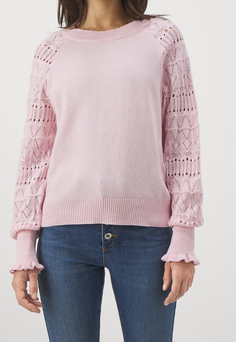 Pull en maille rose clair avec manches longues semi-transparentes à motifs et poignets à volants, porté avec un jean en denim bleu.
