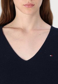 Gros plan sur le bas du visage et le haut de la poitrine d'une femme portant un pull côtelé bleu marine à col en V avec un petit logo drapeau sur le côté droit.