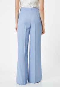 Pantaloni ampi a vita alta di colore azzurro chiaro, con una texture liscia e una parte frontale plissettata. Presentano una chiusura con zip e non hanno tasche visibili.