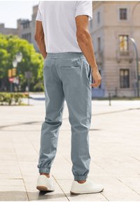 Lichtgrijze joggers met elastische boorden en achterzakken, gecombineerd met witte sneakers, gedragen door een persoon die met de rug naar ons staat, buiten.