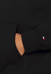 Svart sweatshirt med en framficka och ribbade ärmavslut. Har en liten röd, vit och blå logotyp på ärmen. Tillverkad av mjukt tyg.