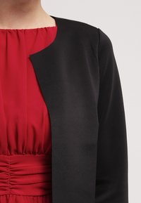 Zwarte blazer met een ronde halslijn, een gladde textuur, open voorkant, gecombineerd met een rode, gathered jurk met een hoge halslijn en geplooid ontwerp.