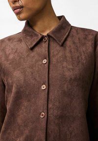 Brun mockasinskjorta med spetsig krage, fem knappar och en mjuk struktur. Har plastknappar i matchande brun ton.