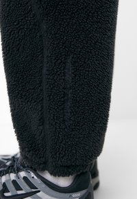 Svart fleecebyxa med en texturerad, fluffig yta och synlig sömnad. Grå sportskor med svarta accenter och vit sula.