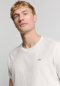 Witte T-shirt met korte mouwen, gemaakt van zacht materiaal, met een klein "NXS"-logo op de borst in lichte en donkere kleuren. Eenvoudig ontwerp.