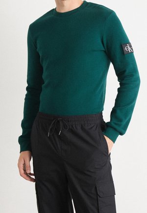 Pull vert côtelé à manches longues, col rond, avec un patch logo noir sur la manche gauche, associé à un pantalon noir à cordon de serrage.
