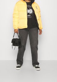 Giacca puffer gialla, t-shirt nera con grafica, jeans grigio scuro e borsa nera con un design testurizzato. Sneakers nere con dettagli bianchi.