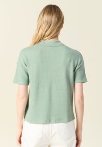 Top a maniche corte con colletto in tessuto verde chiaro testurizzato. Presenta una vestibilità rilassata e un orlo arrotondato. La vista posteriore mostra un colletto liscio.