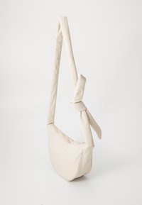 Beige tekstil skulderveske med en knute-detalj på stroppen, med rund form og glatt tekstur. Glidelås lukking på toppen.