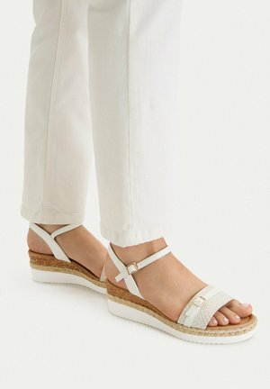 Pies con sandalias blancas de cuña con tiras, corcho y suelas blancas, combinadas con pantalones blancos de pierna recta.