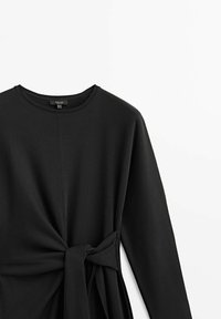 Haut noir à manches longues en tissu doux, présentant un col rond et un détail noué à la taille, créant un effet froncé.