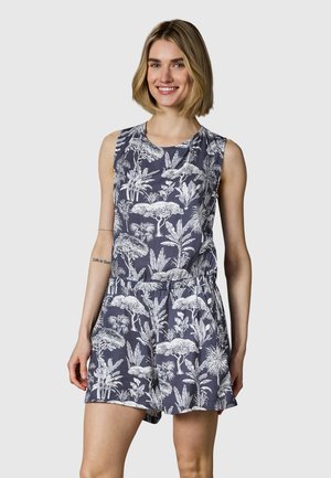 Uśmiechnięta kobieta ubrana w granatowy kombinezon bez rękawów z białym tropikalnym wzorem drzew i roślin, stojąca na tle jednolitego jasnego tła.