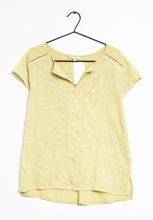 Blouse jaune pâle à manches courtes avec motif de feuilles texturé, encolure ouverte et étiquette en tissu, suspendue à un cintre noir sur fond blanc.