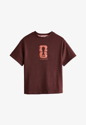 Ung pige med langt hår iført en brun FIFA World Cup t-shirt, vidde jeans og sneakers, lænende mod en beige væg.