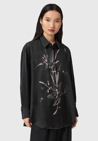 Femme portant une chemise noire ample à boutons avec un grand motif floral gris sur le devant, se tenant devant un fond neutre.