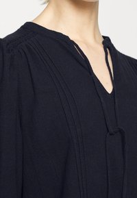 Blouse bleu marine avec des détails plissés aux épaules et un décolleté en V. Fabriquée en tissu doux avec une coupe décontractée et un accent de nœud visible.