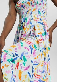 Donna che indossa un vestito senza maniche con un corpetto arricciato, sollevando la gonna per rivelare un motivo colorato di pennellate astratte su un tessuto bianco.