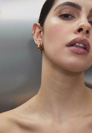 Close-up af en kvindes ansigt og hals, der viser glat hud, blanke læber, mørke øjne og guldøreringe på en neutral baggrund.
