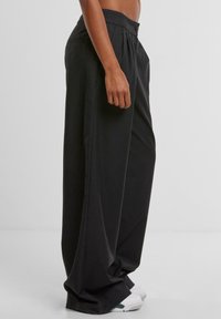 Personne debout de profil, portant un pantalon noir ample et des baskets blanches, le ventre nu sur un fond clair et uni.