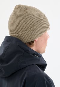 Henkilö, jolla on päällään beige neulottu pipo ja musta huppari, katsoo oikealle yksinkertaista vaaleaa taustaa vasten.