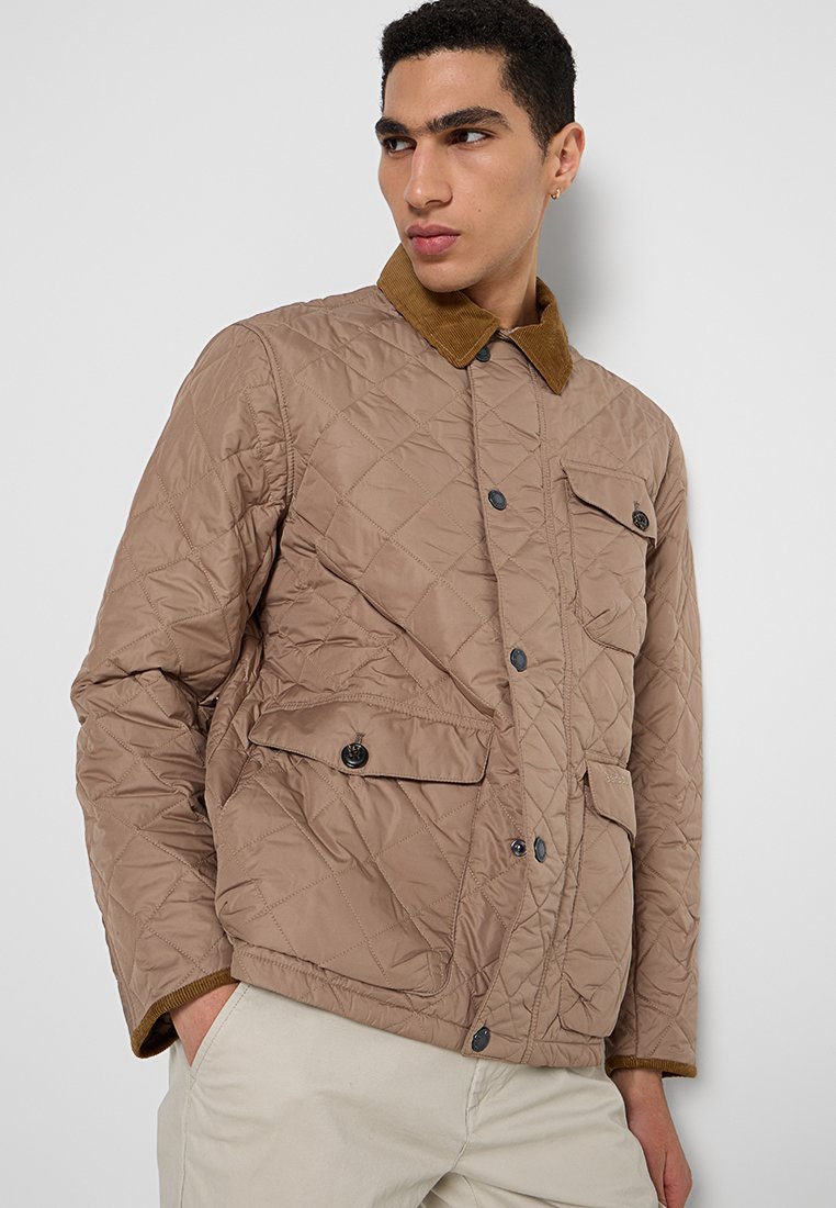 Homme portant une veste matelassée marron clair avec des poches à rabat et un col en velours côtelé contrasté, les mains dans les poches, regardant sur le côté.