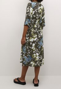 Florales Midikleid in Olivgrün mit blauen und weißen Blumen. Lockerer Schnitt mit kurzen Ärmeln und tailliertem Gürtel. Kombiniert mit schwarzen Sandalen.