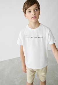 Valkoinen puuvillapaita, jossa on musta teksti "BAKER BY TED BAKER". Yhdistetty vaaleanbeigeihin shortseihin, neutraalilla taustalla.