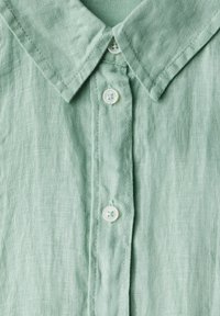 Camicia di lino verde chiaro con collo alla coreana, patta con tre bottoni bianchi e superficie in tessuto testurizzato.