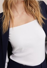 Bílý žebrovaný top na ramínka se čtvercovým výstřihem, zkombinovaný s tmavě modrým blejzrem. Tkanina vypadá měkce a přiléhavě.