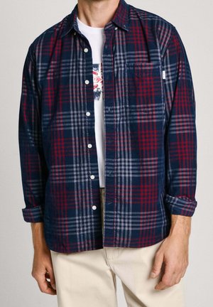 Muž ve tmavě modré, červené a šedé kárované košili s knoflíky přes bílým tričkem s potiskem, s vyhrnutými rukávy a béžovými kalhotami.