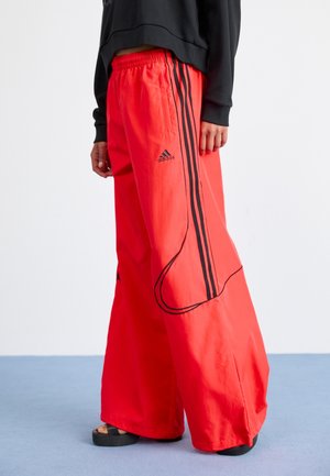 Personne portant un pantalon large Adidas rouge vif avec des bandes noires et un logo, associé à un haut noir à manches longues et des sandales noires.