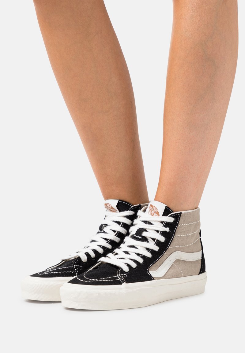 Hög-top sneakers med en svart kanvasutsida, beige detaljer, vita snören och en vit gummisula. Anmärkningsvärd logotyp på sidan.