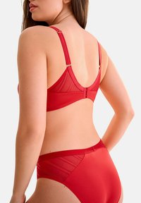 Ensemble lingerie rouge en dentelle comprenant un soutien-gorge et une culotte, présentant des panneaux transparents, des bretelles réglables et des accents en doré, avec une texture lisse et une coupe ajustée.