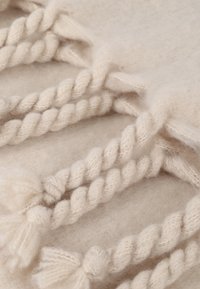 Cremefarvet tæppe med en blød uldtekstur, prydet med snoede rebdetaljer og pom-pom detaljer for ekstra visuel interesse.