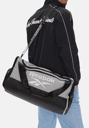 Muž v černé bundě a světle modrých džínách nese přes rameno šedou a černou tašku Reebok.