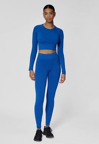 Niebieski zestaw sportowy składający się z długiego, krótko skrojonego topu z długim rękawem i legginsów z wysokim stanem o teksturowanym wzorze, połączony z czarnymi butami sportowymi.