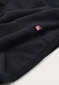Schwarzer Fleece-Stoff mit glatter Textur, auf der Seite mit einem roten und schwarzen Etikett genäht, das das "66° North"-Logo zeigt.