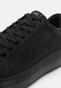 Juodi sportbačiai su zomšos viršumi, tekstūruotais akcentais, apvaliais raišteliais ir blizgia juoda stora semtimi. Pusėje iškilmingai įspaustas "KARL" logotipas.