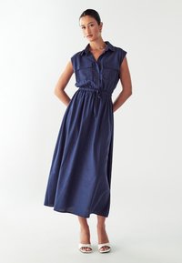 Abito senza maniche di colore blu navy con chiusura a bottoni sul davanti, due tasche sul petto, vita elasticizzata e gonna fluida, abbinato a tacchi bianchi.