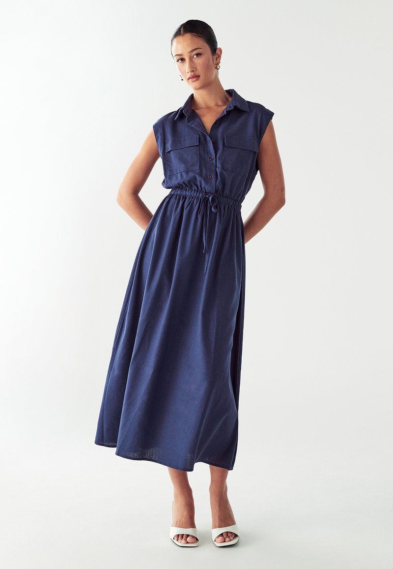 Abito senza maniche di colore blu navy con chiusura a bottoni sul davanti, due tasche sul petto, vita elasticizzata e gonna fluida, abbinato a tacchi bianchi.