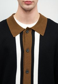 Férfi fekete kötött pólót visel barna gallérral, középen függőleges barna, fekete és fehér csíkokkal, fekete gombokkal.