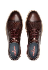 Bruine leren schoenen met een gestructureerde mesh bovenkant, contrasterende bordeauxrode veters en een lichtbruine zool. Merkenlogo zichtbaar op de binnenzool.