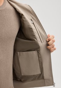 Beżowa podszewka kurtki z gładkiego materiału, kieszeń na zamek oraz dodatkowa płaska kieszeń. Zewnętrzna część ma czysty, elegancki design.