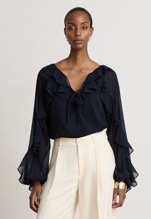 Femme portant un chemisier noir à manches longues avec volants et nœud au col ainsi qu’un pantalon blanc taille haute sur un fond uni.