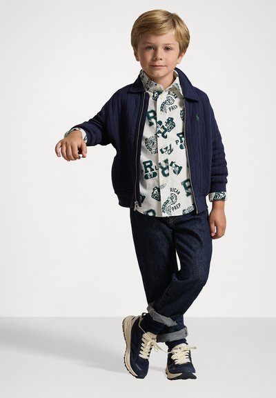 Casaco acolchoado azul-marinho, camisa branca com gráficos impressos azuis, jeans de denim escuro e ténis azul-marinho com atacadores creme. Pose em pé com os braços relaxados.