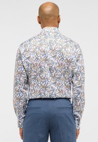 Camicia bianca a maniche lunghe con un colorato motivo paisley in verde, blu e arancione; abbinata a pantaloni blu.